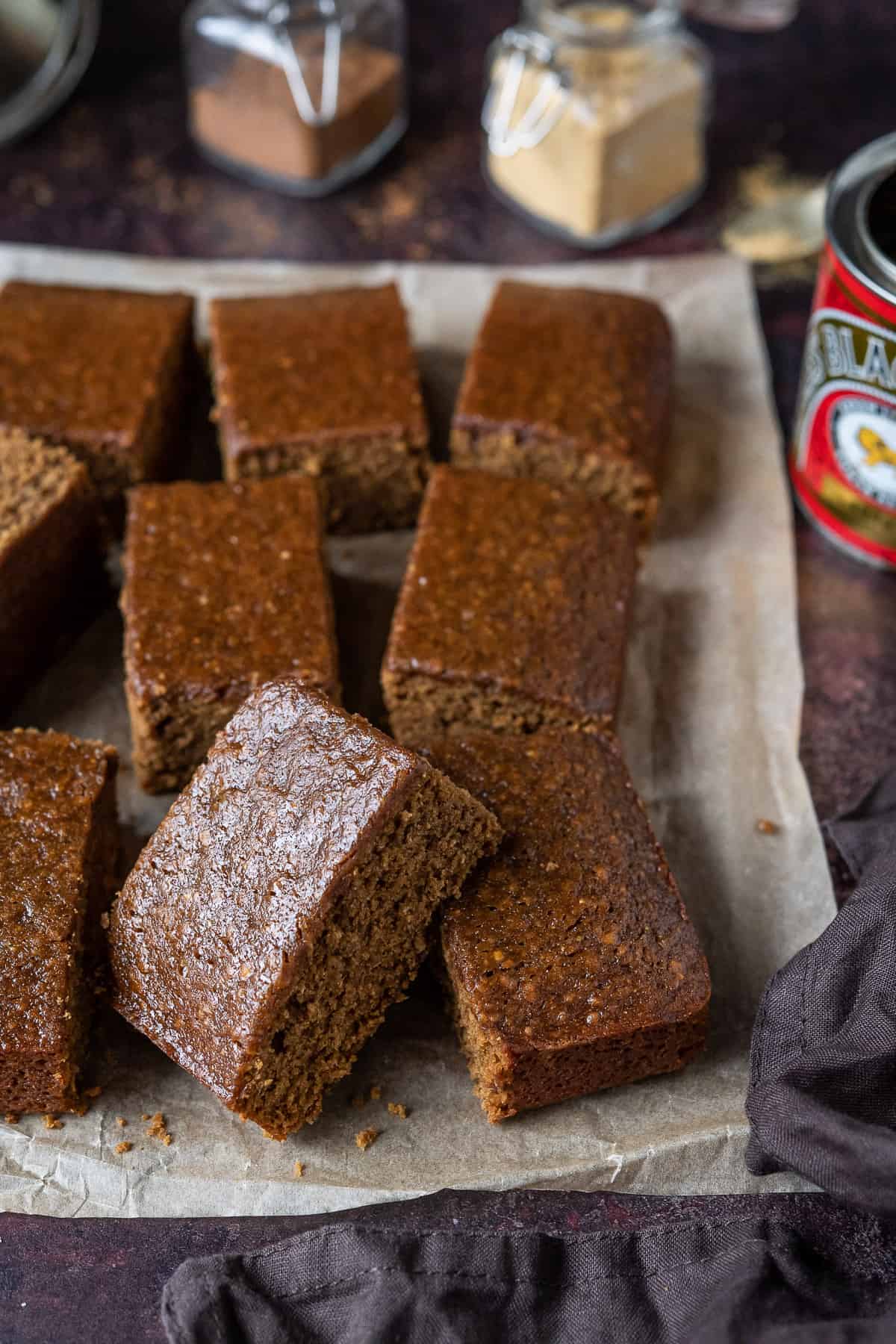 Squares of vegan parkin on brown parchment on a dark brown background with a tin of treacle and jars of spices.