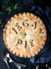 Chickpea, leek and mushroom pie with the words "vegan pie" written on top, on a dark background with forks, parsley, salt and pepper shakers and a grey cloth.
