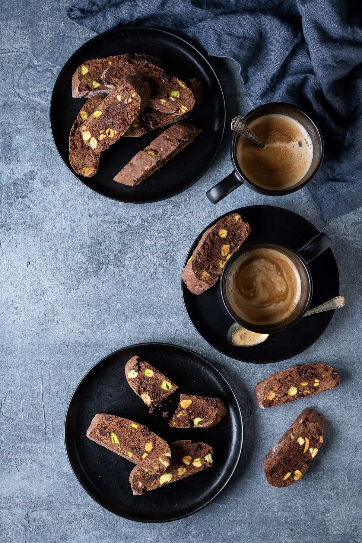 Chocolate pistachio biscotti on black plates with black cups of espresso coffee and a grey cloth.