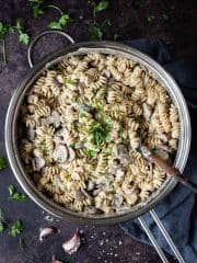 A metal pan of creamy vegan mushroom pasta on a dark surface with fresh parsley and garlic skins.