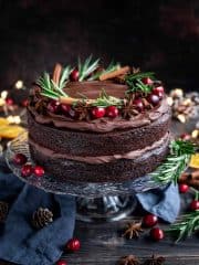 Vegan mulled wine chocolate cake topped with cranberries, rosemary, star anise and cinnamon sticks on a glass cake stand on a wooden table.