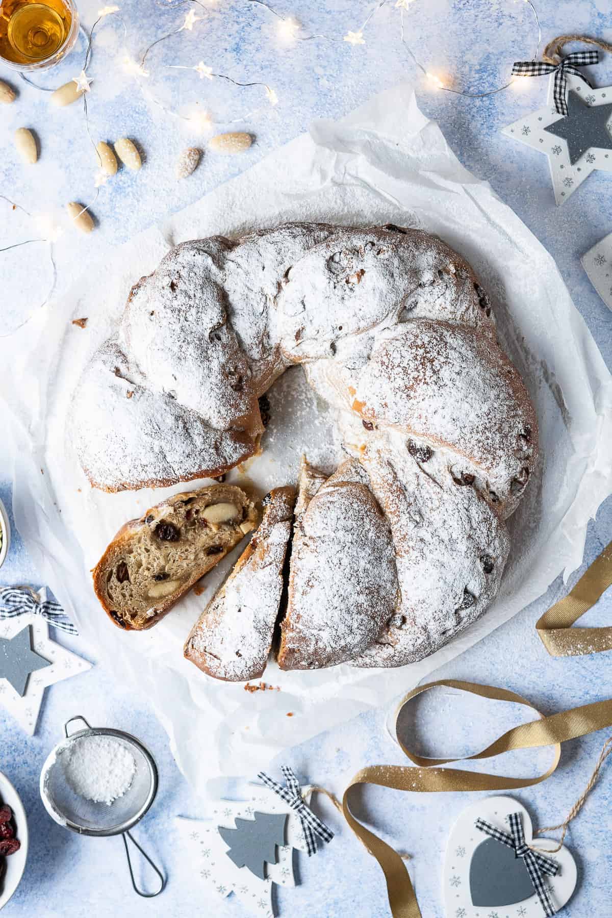 Vegan stollen wreath on a sheet of white baking parchment on a blue surface with christmas decorations and lights.