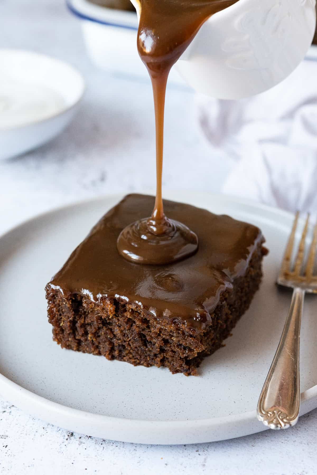 A square of vegan sticky toffee pudding on a grey plate with toffee sauce being poured on top.