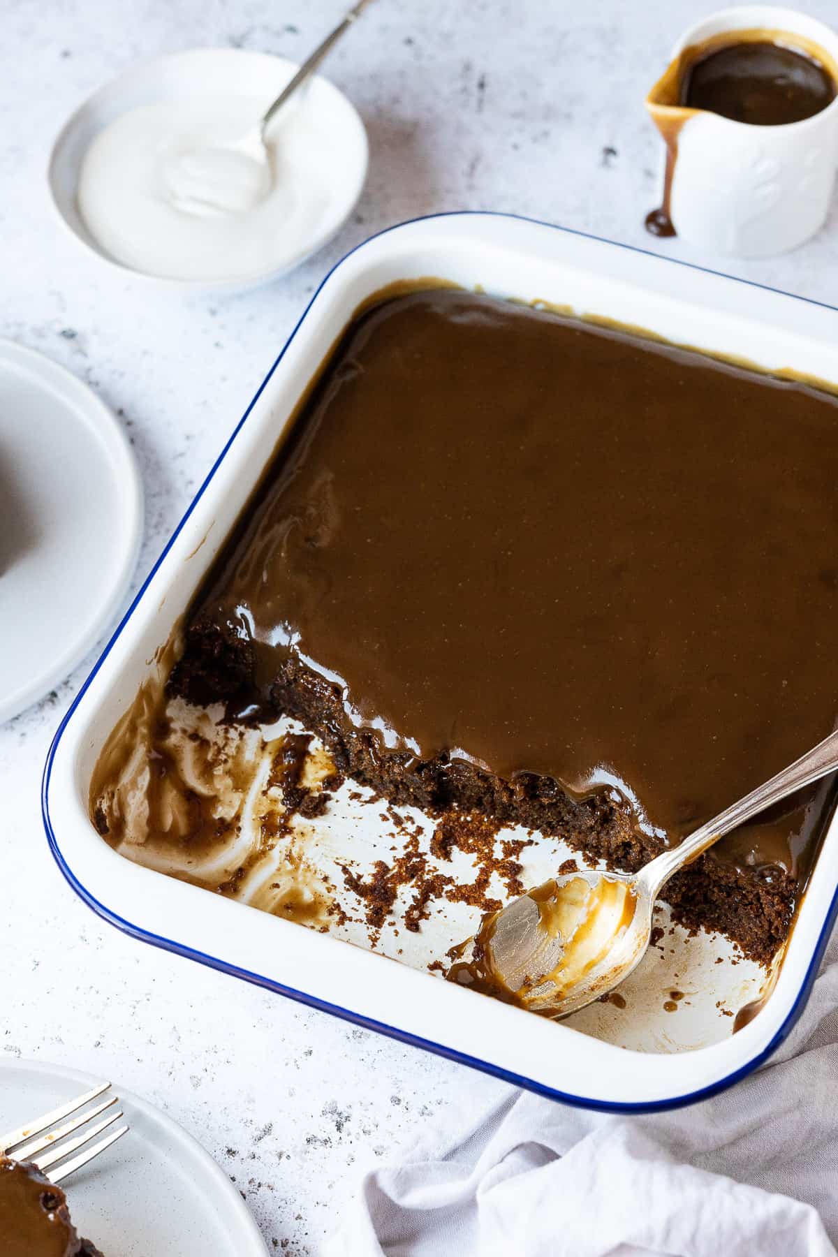 Sticky toffee pudding in a white enamel dish on a white background with a jug of sauce and a bowl of cream.