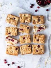 Vegan almond cranberry cake sliced into bars on a sheet of baking parchment on a white background with a bowl of fresh cranberries.