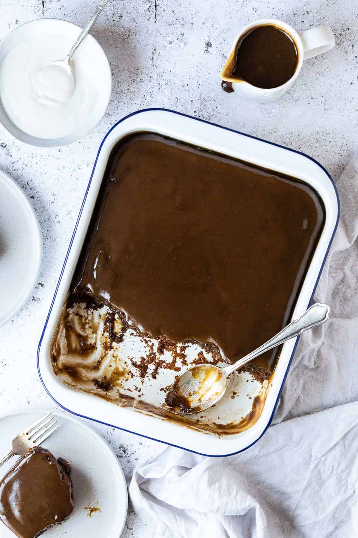 Vegan sticky toffee pudding in an enamel baking dish with several portions scooped out onto plates, with a jug of toffee sauce and a bowl of cream.