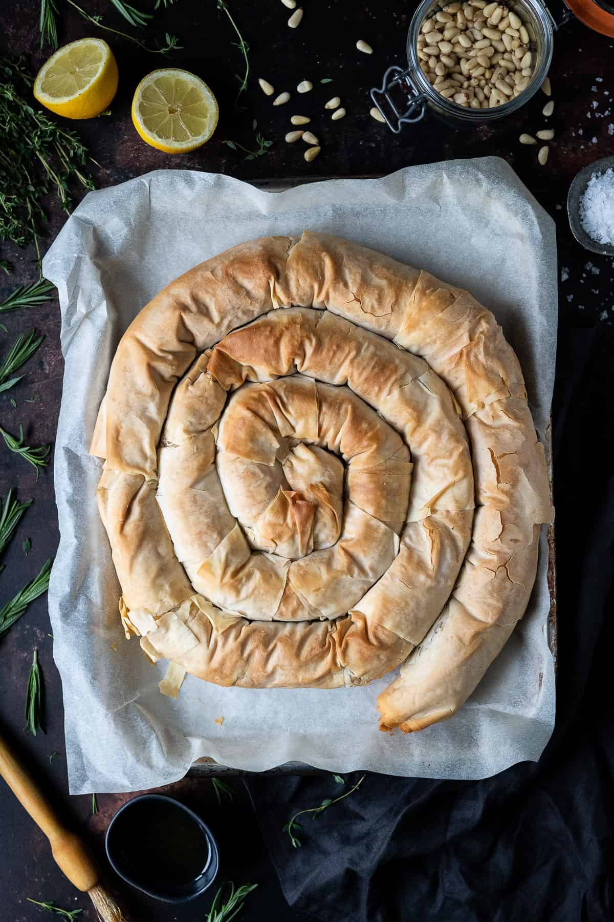 Vegan butternut squash and feta spiral pie on a baking parchment lined baking sheet on a dark background surrounded by fresh herbs, a jar of pine nuts, a halved lemon, pastry brush and bowl of olive oil.