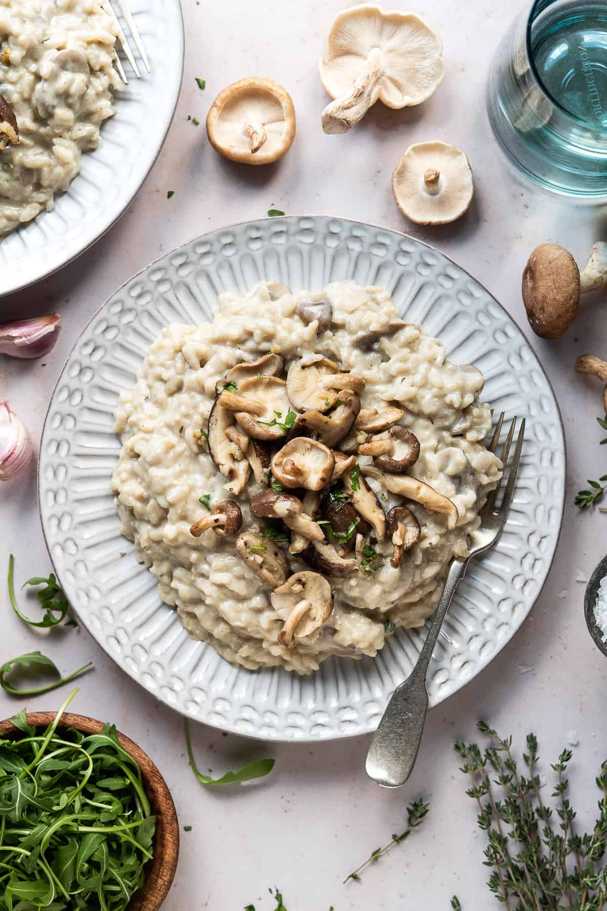 A plate of vegan mushroom risotto surrounded by a bowl of rocket, fresh thyme, garlic skins, a glass of water and some raw mushrooms.