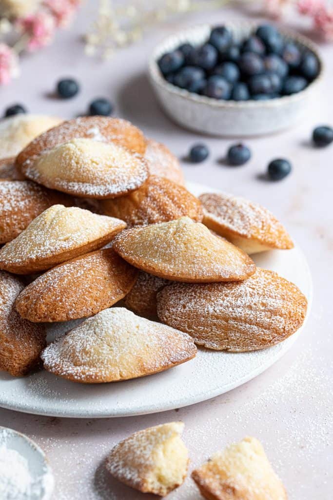 A pile of Madeleines on a white plate with a bowl of blueberries.