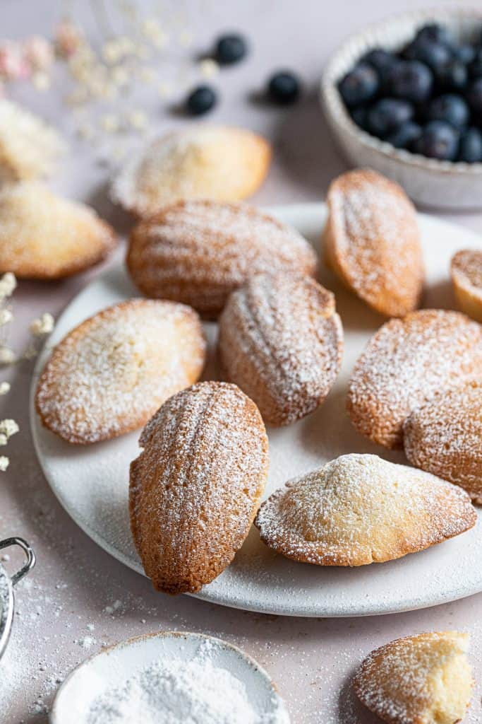 Vegan Madeleines on a white plate with a bowl of blueberries in the background.