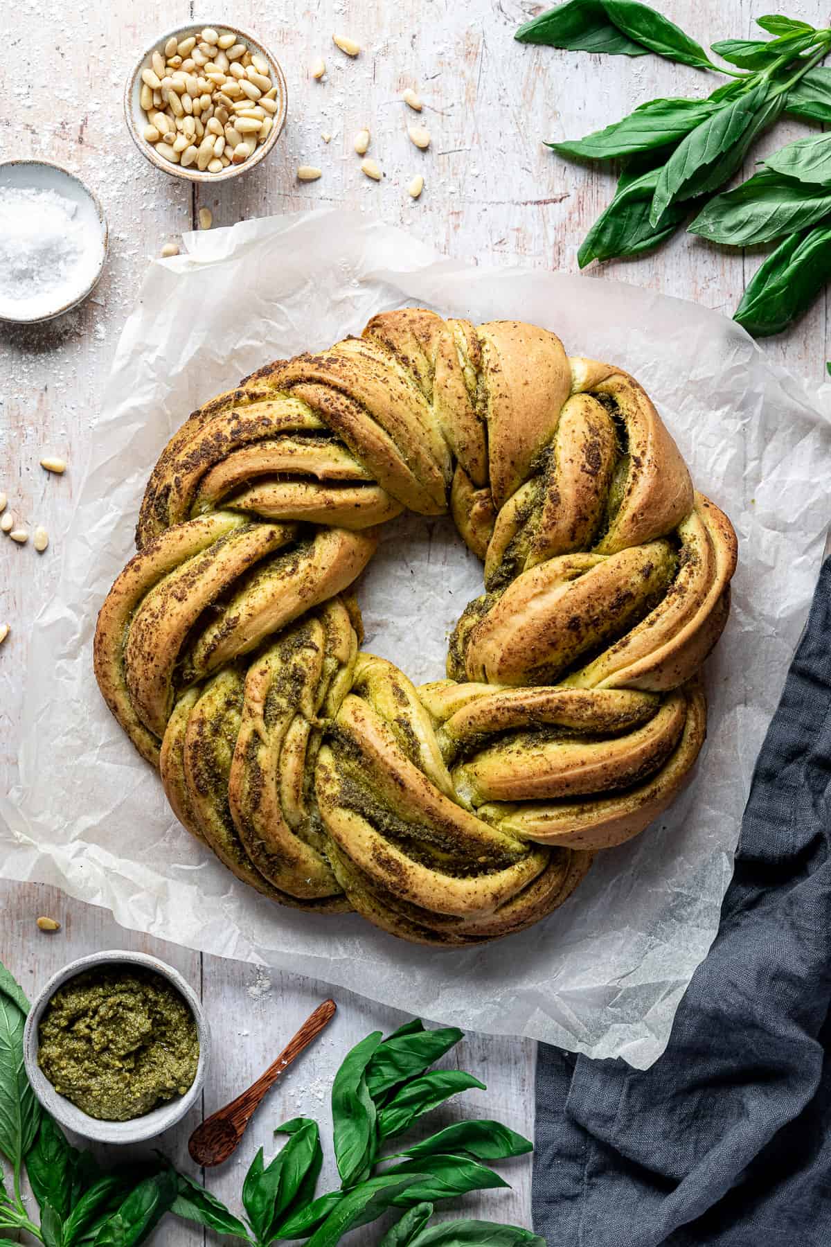 Vegan pesto bread wreath on a sheet of baking parchment on a wooden table surrounded by fresh basil and bowls of pine nuts, salt and pesto.