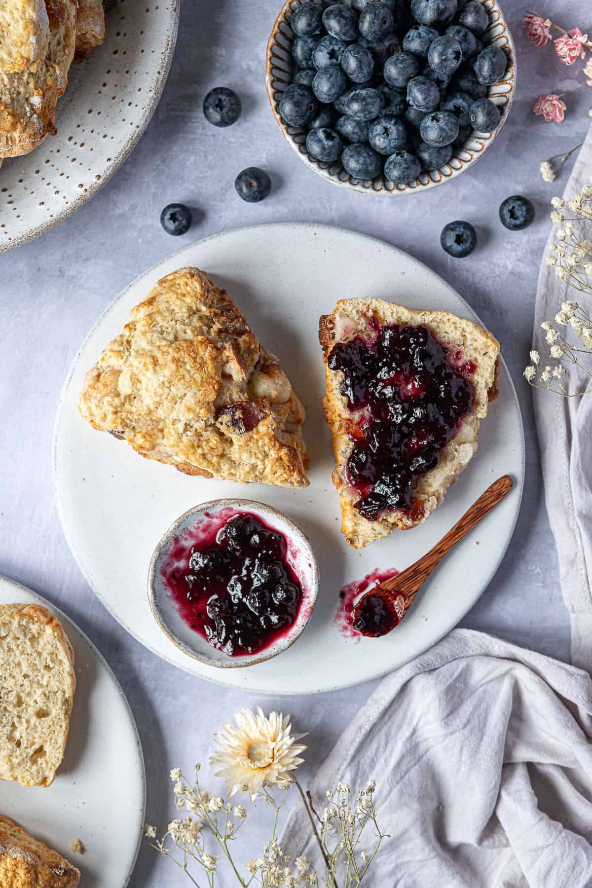 A scone sliced in half on a white plate with jam, surrounded by a bowl of blueberries, dried flowers, grey linen and plates of scones.