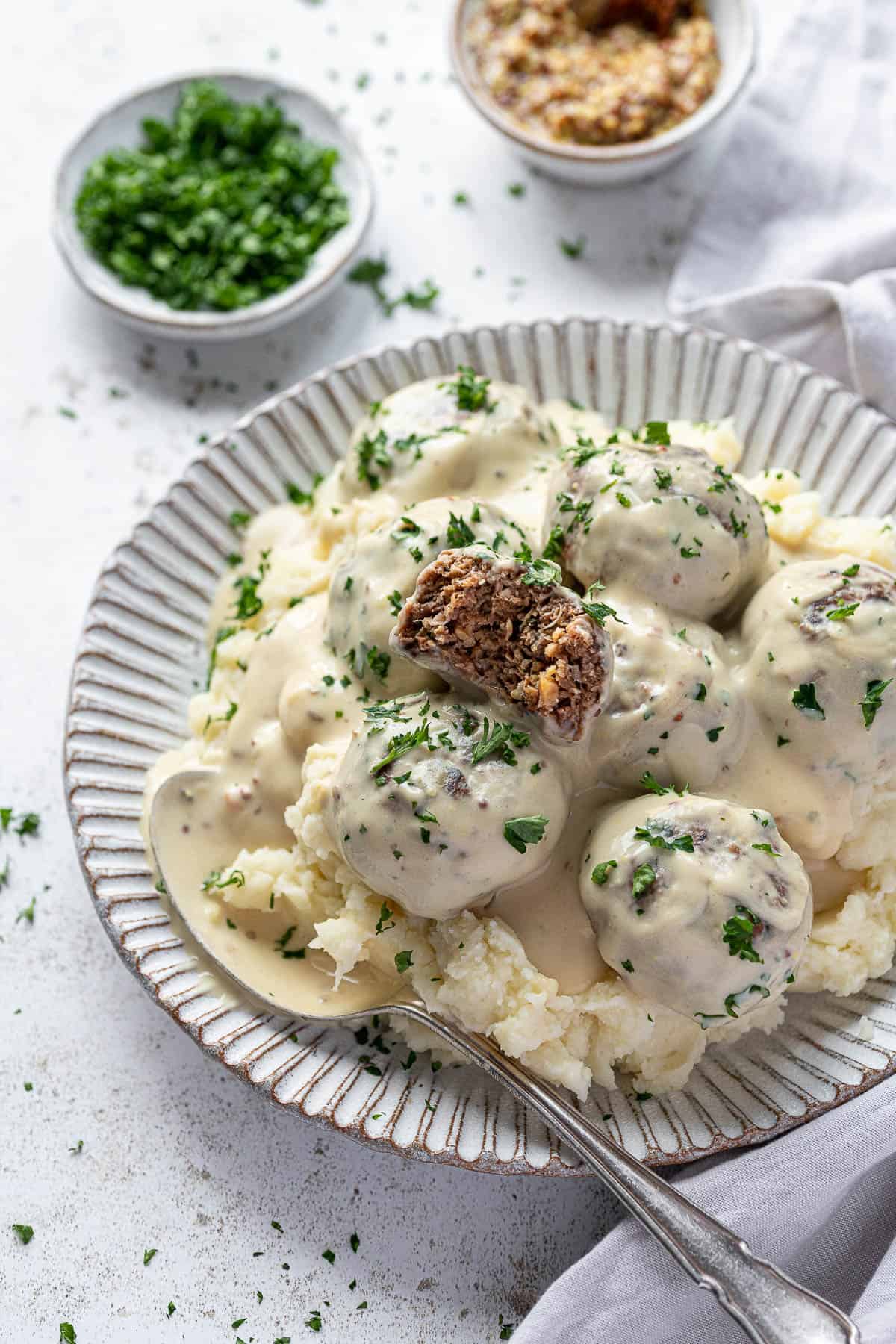 Vegan Swedish meatballs and mashed potato on a white plate with a bowl of parsley and a bowl of mustard.