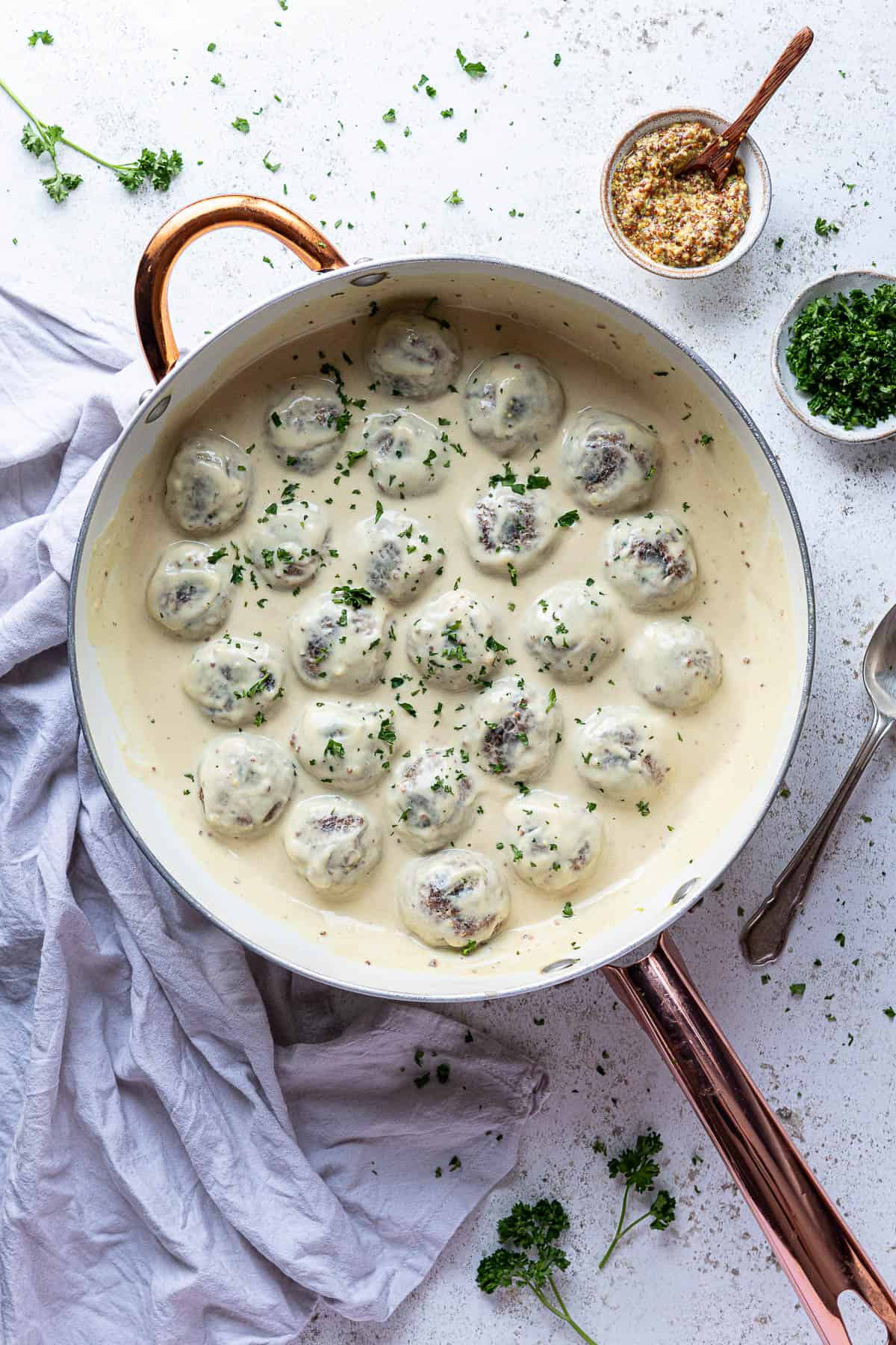 Vegan Swedish meatballs in a creamy mustard sauce in a white saucepan with a bowl of mustard and a bowl of chopped parsley.