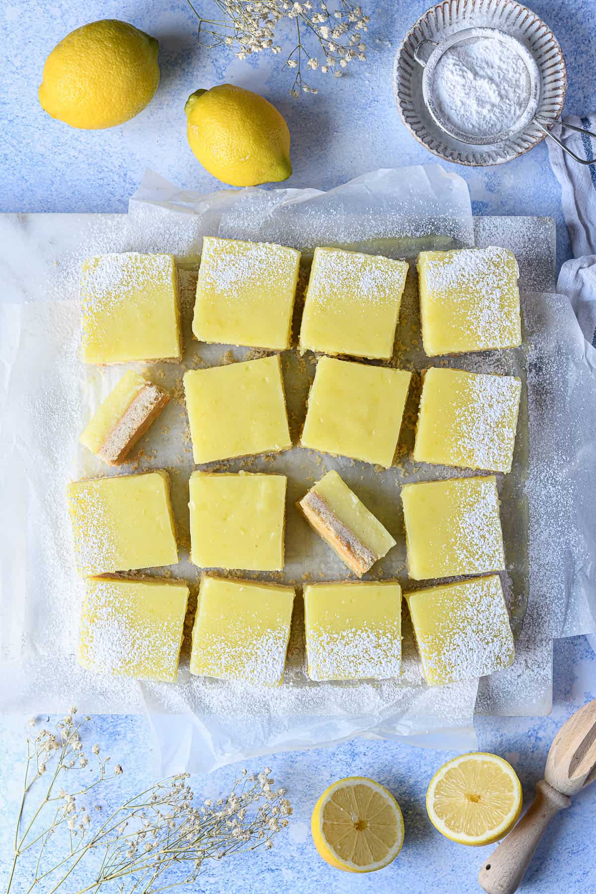 Vegan lemon bars on a blue surface with fresh lemons, a bowl of icing sugar and dried flowers.