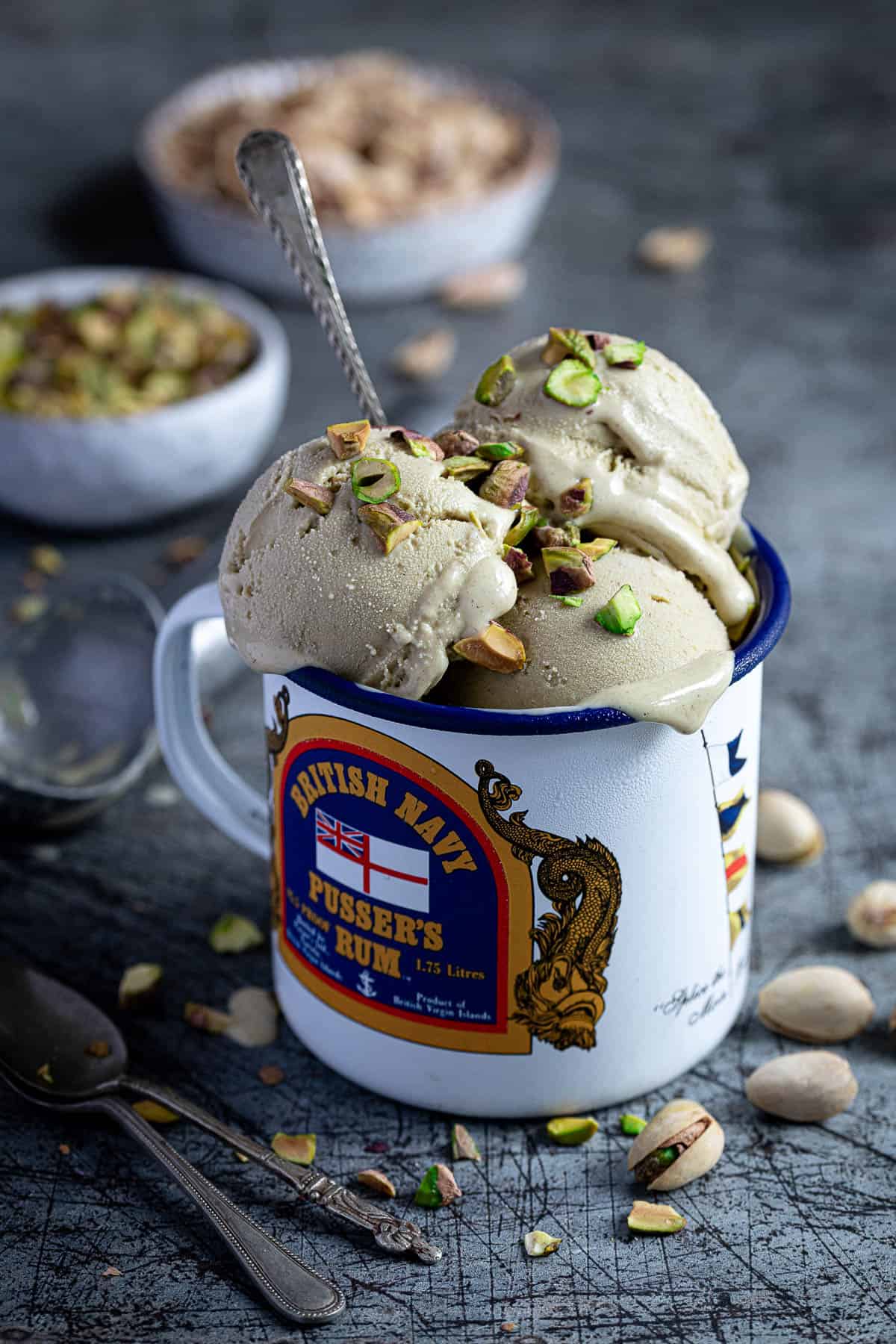 Scoops of vegan pistachio ice cream in an enamel mug with a bowl of chopped pistachios and a bowl of pistachio shells.