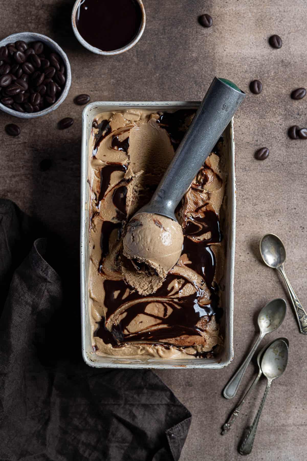 Vegan coffee ice cream in a metal tin on a brown surface with a bowl of chocolate sauce, bowl of chocolate coffee beans, a brown cloth and metal tea spoons.
