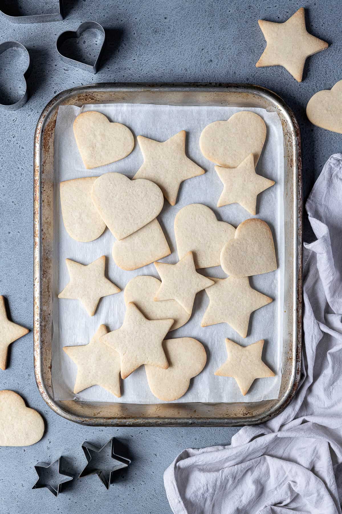 Vegan vanilla sugar cookies on a meatl baking tray with cookie cutters and a grey cloth.