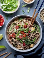 Creamy peanut noodles with stir fried vegetables in a bowl with chopsticks and bowls of spring onions, peanut butter and chillies.
