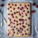 Vegan cherry almond cake on a sheet of baking parchment with bowls of cherries and flaked almonds.
