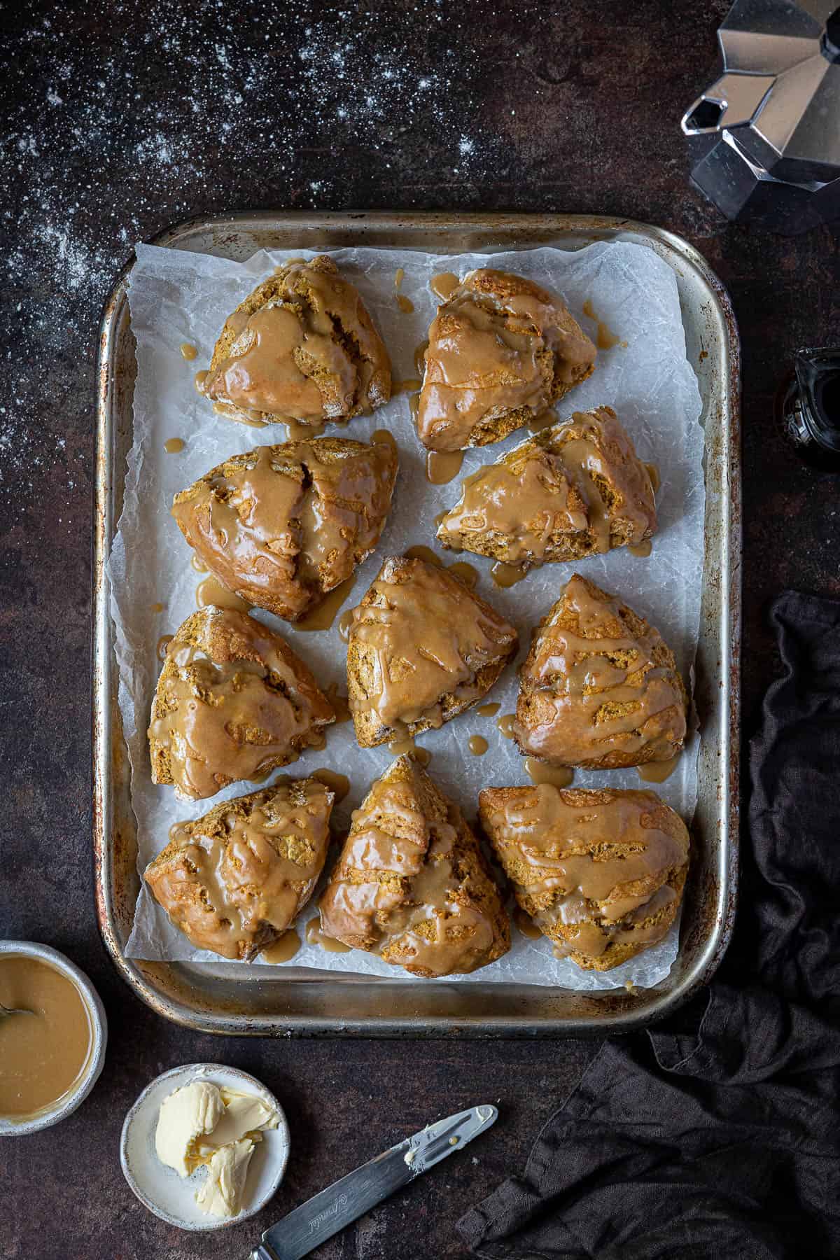 Vegan pumpkin scones on a metal baking tray with a coffee pot, bowl of maple glaze and dish of butter.