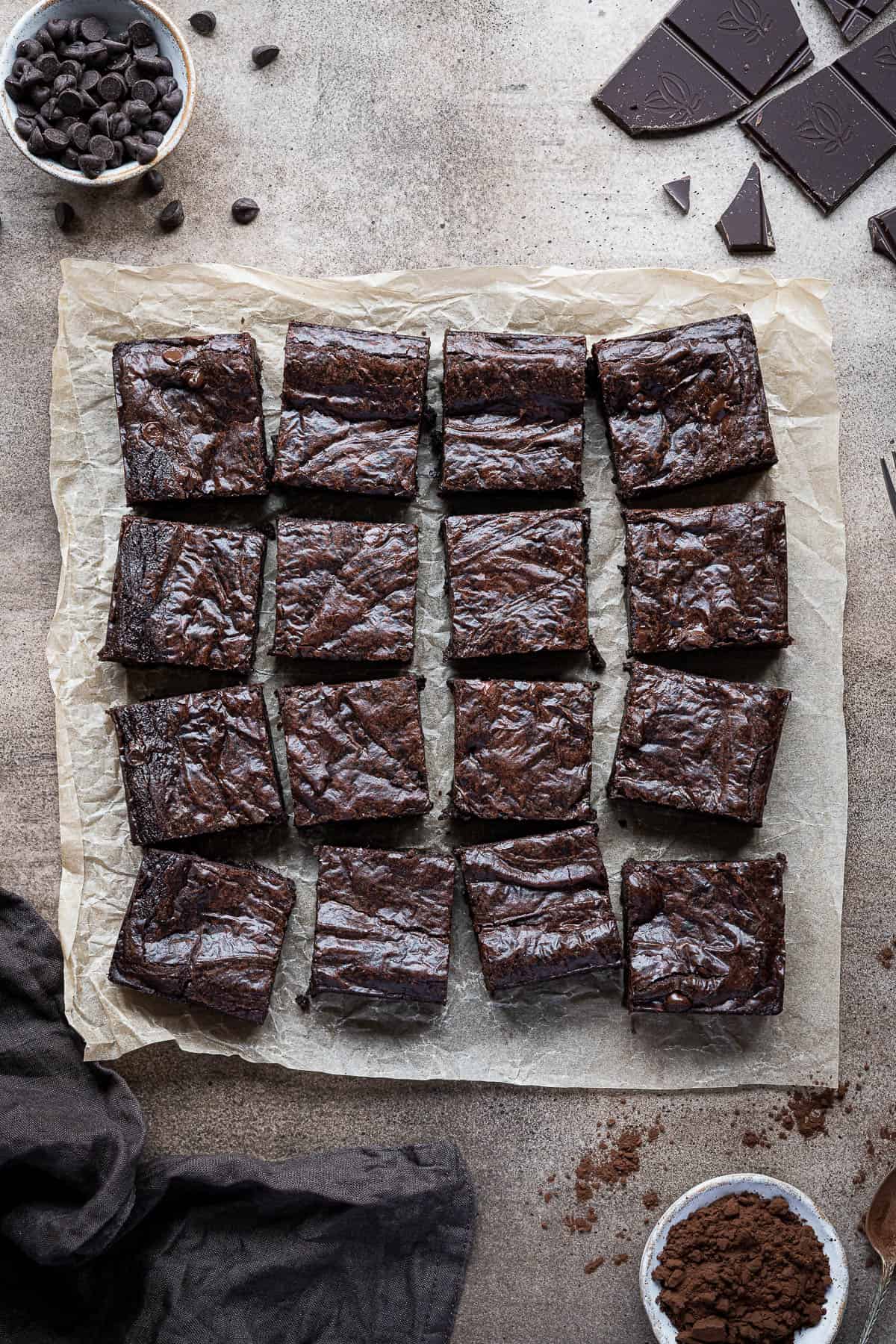 Fudgy vegan brownies on a sheet of baking parchment on a brown surface, surrounded by bowls of chocolate chips and cocoa powder, a broken bar of chocolate and a brown cloth.
