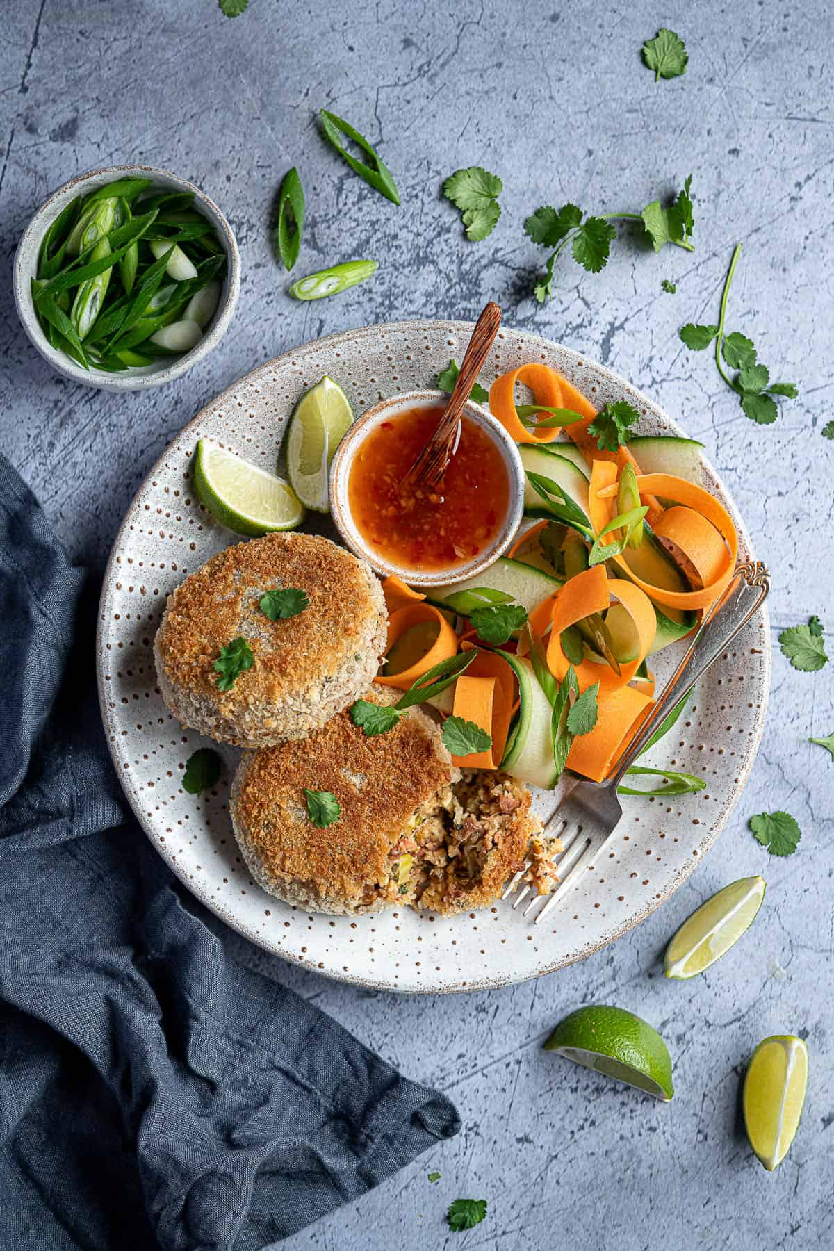 Two vegan Thai red curry fish cakes on a plate with vegetable ribbons and a bowl of sweet chilli sauce, surrounded by lime slices, sliced spring onions and fresh coriander.