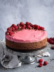 Vegan chocolate raspberry mousse cake on a glass cake stand with a grey cloth and fresh raspberries.