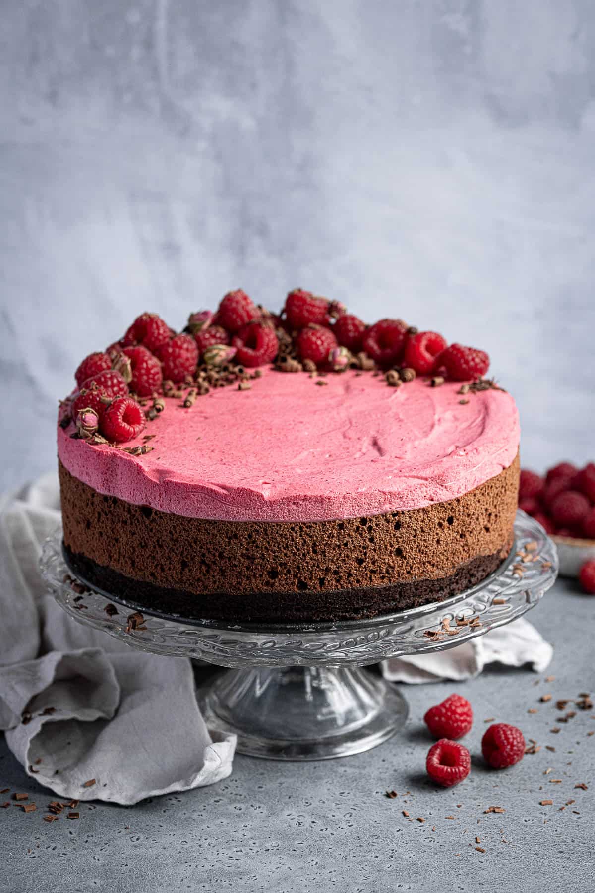 Vegan chocolate raspberry mousse cake on a glass cake stand with a grey cloth and fresh raspberries.