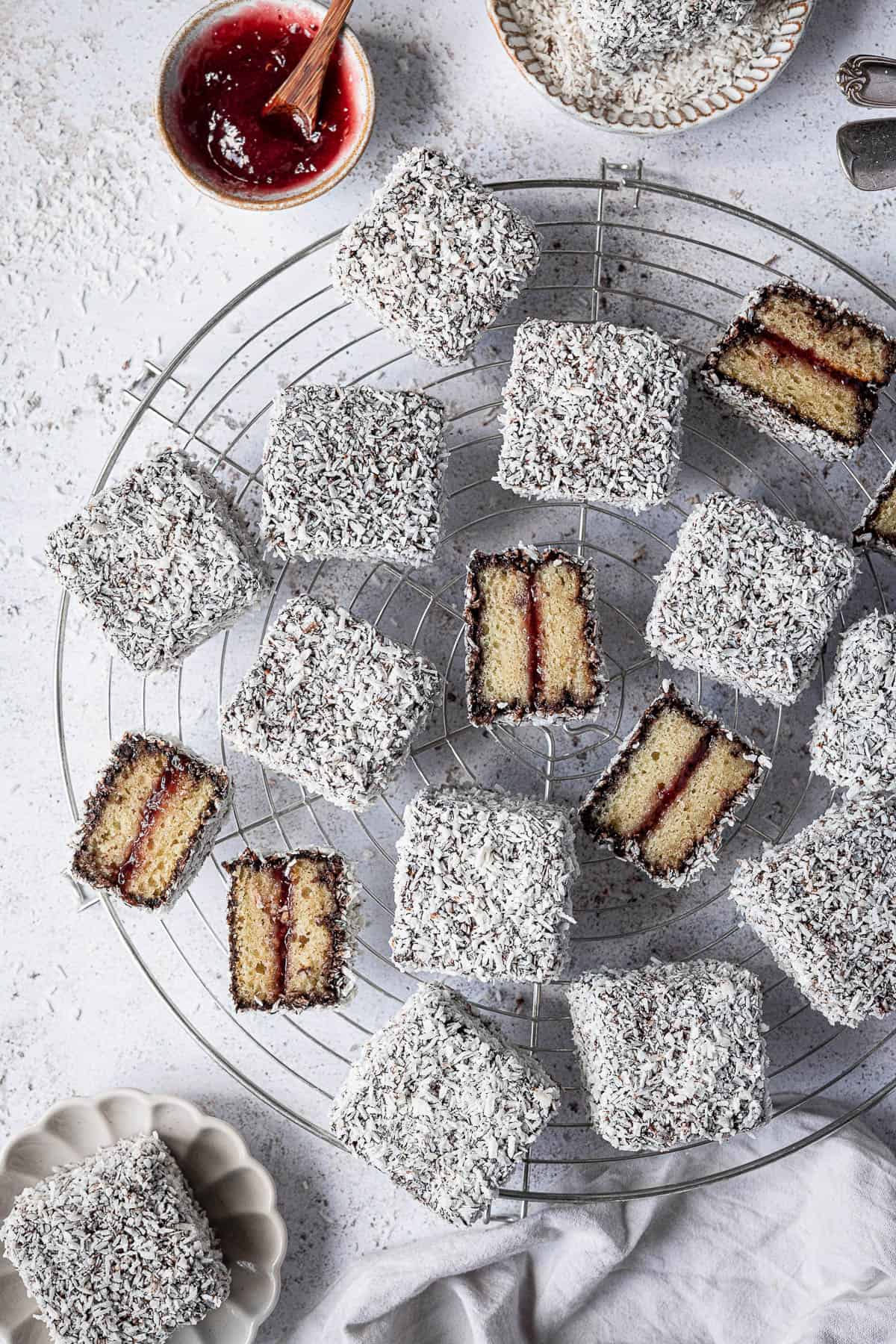 Vegan Lamingtons on a round wire rack with a bowl of jam, some sliced in half.