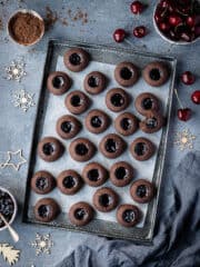 Vegan chocolate cherry thumbprint cookies on a metal tray surrounded by wooden Christmas ornaments and bowls of cherries, jam and cocoa powder.