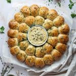 Vegan garlic dough ball wreath on a sheet of baking parchment surrounded by bowls of herbs and garlic butter.