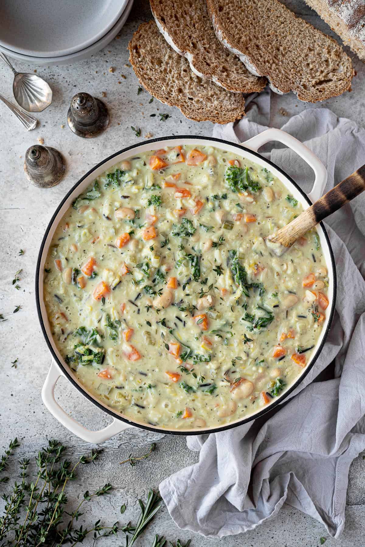 A pan of creamy vegetable rice soup with slices of bread, fresh herbs, soup bowls and salt and pepper shakers.