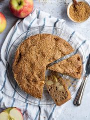 Simple wholemeal apple cake with two slices cut out on a blue and white cloth surrounded by apples and a bowl of sugar.