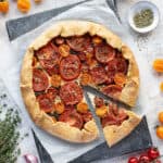 Vegan tomato ricotta galette with a slice cut out on a sheet of baking parchment surrounded by tomatoes, herbs and a bowl of pesto.
