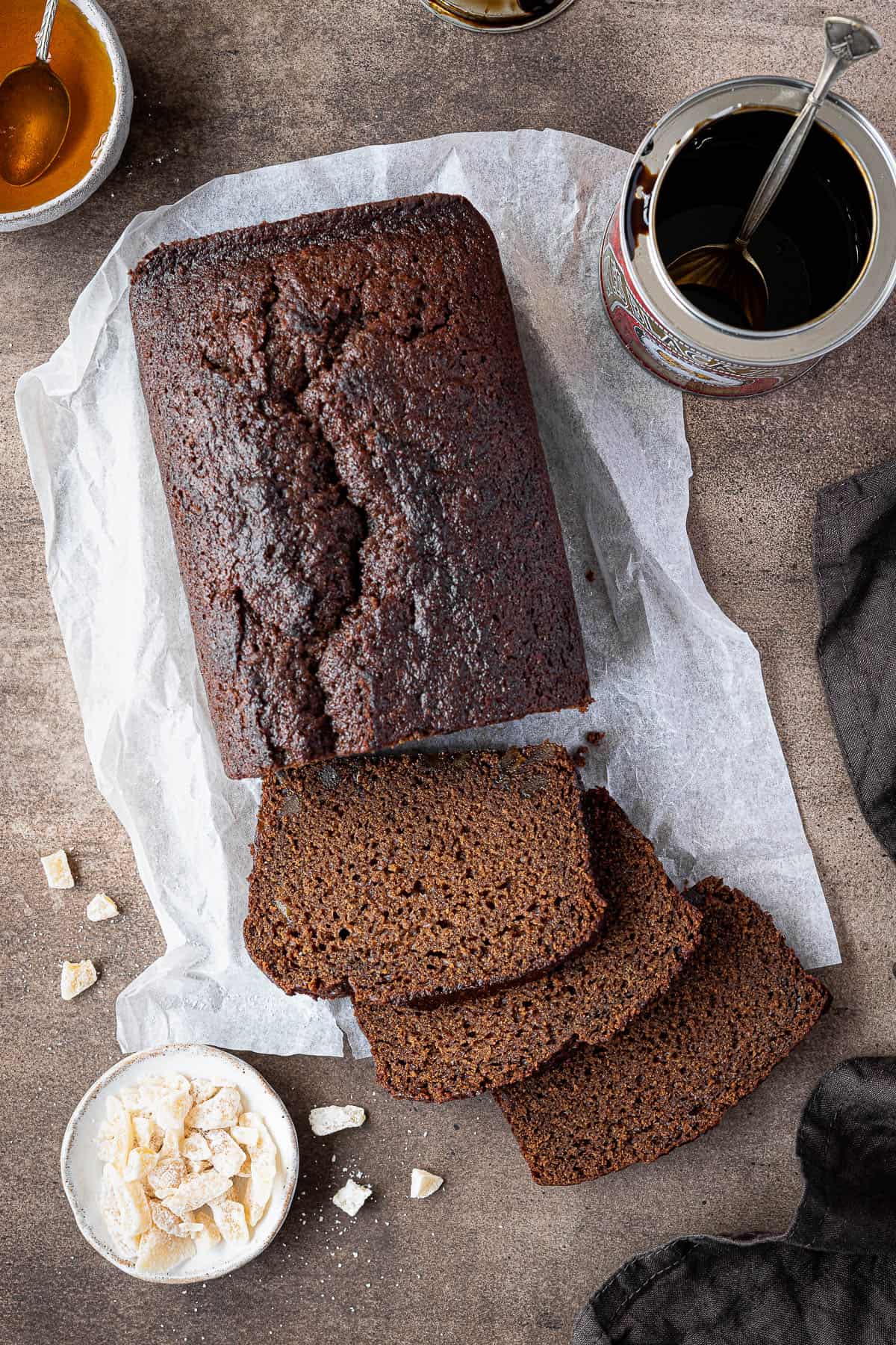 Vegan ginger loaf cake on a brown background with a tin of treacle and bowls of candied ginger and golden syrup.