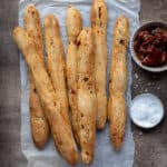 Vegan sun dried tomato breadsticks on a sheet of baking parchment with bowls of sun dried tomatoes and salt.