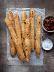 Vegan sun dried tomato breadsticks on a sheet of baking parchment with bowls of sun dried tomatoes and salt.