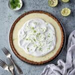 Vegan key lime pie on a grey surface wiith lime halves, a bowl of lime zest and a pile of forks.