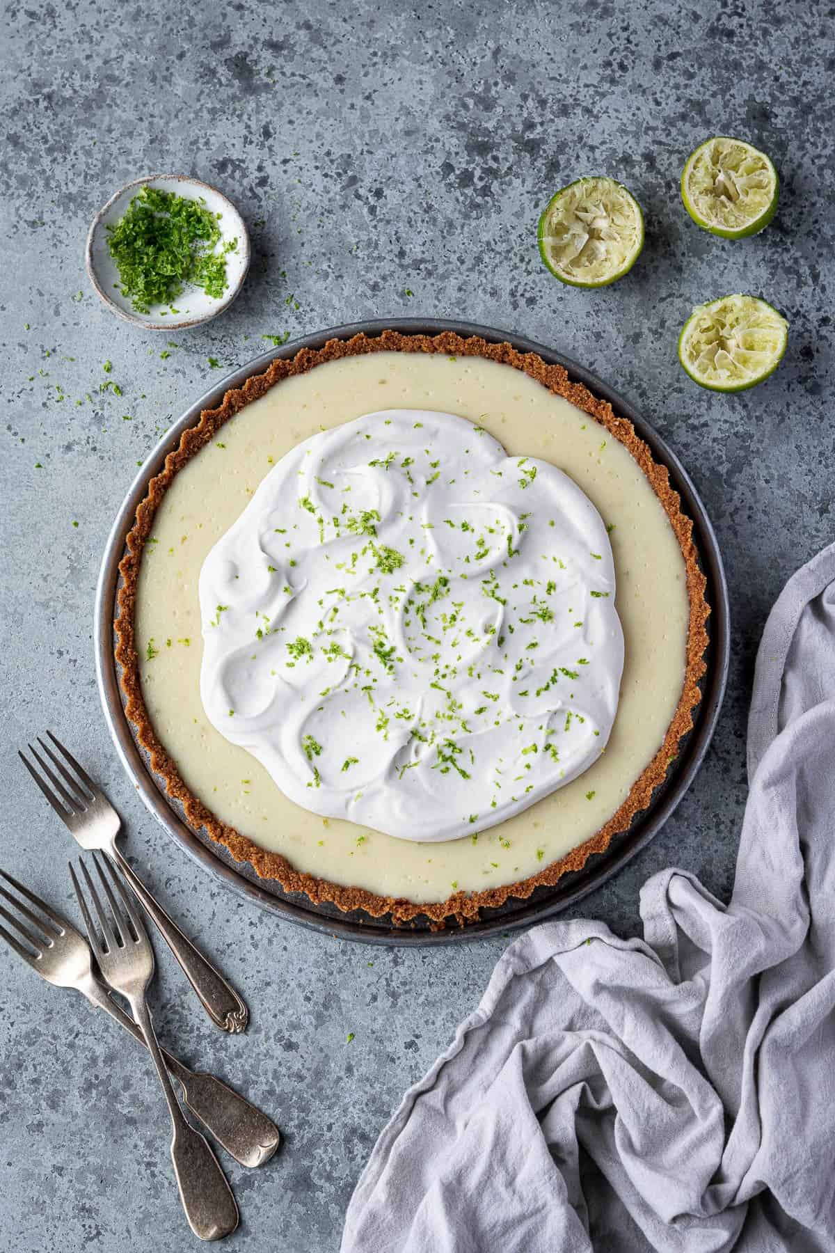 Vegan key lime pie on a grey surface wiith lime halves, a bowl of lime zest and a pile of forks.