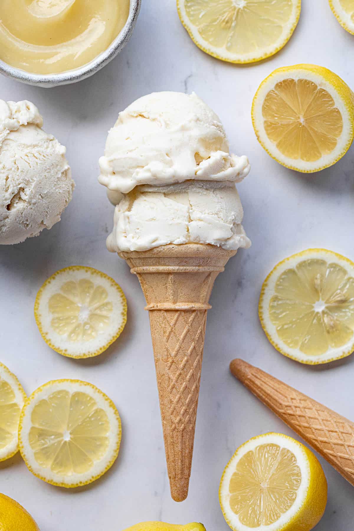 Two scoops of vegan lemon ice cream in a cone, surrounded by lemon slices.