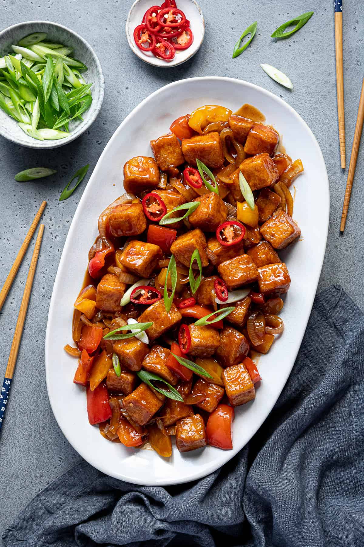 Sweet and sour tofu on an oval plate with chopsticks and bowls of spring onion and chilli.