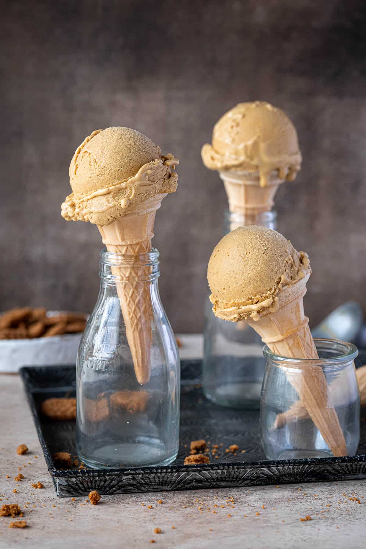 Three cones of vegan pumpkin ice cream in glass milk bottles with a bowl of crushed cookies.