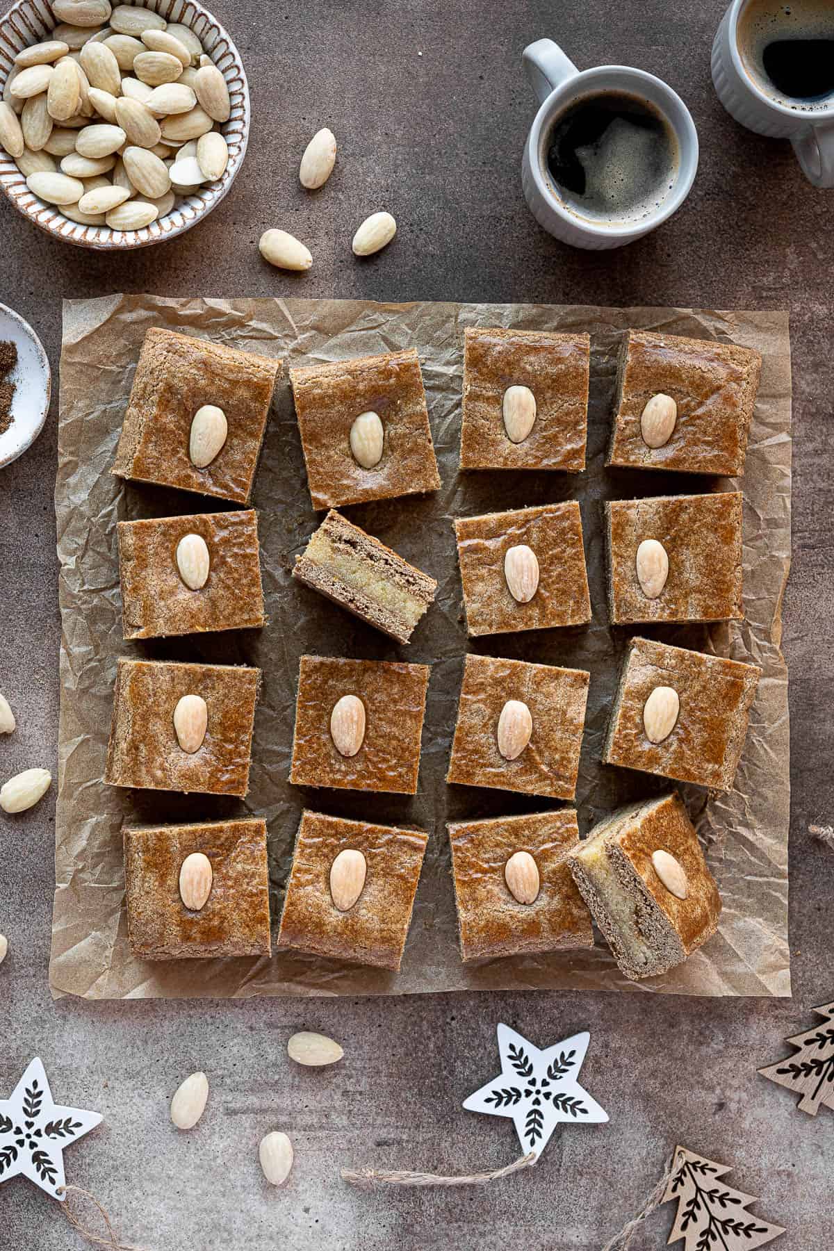 Squares of vegan gevulde speculaas surrounded by cups of coffee, a bowl of almonds and Christmas decorations.