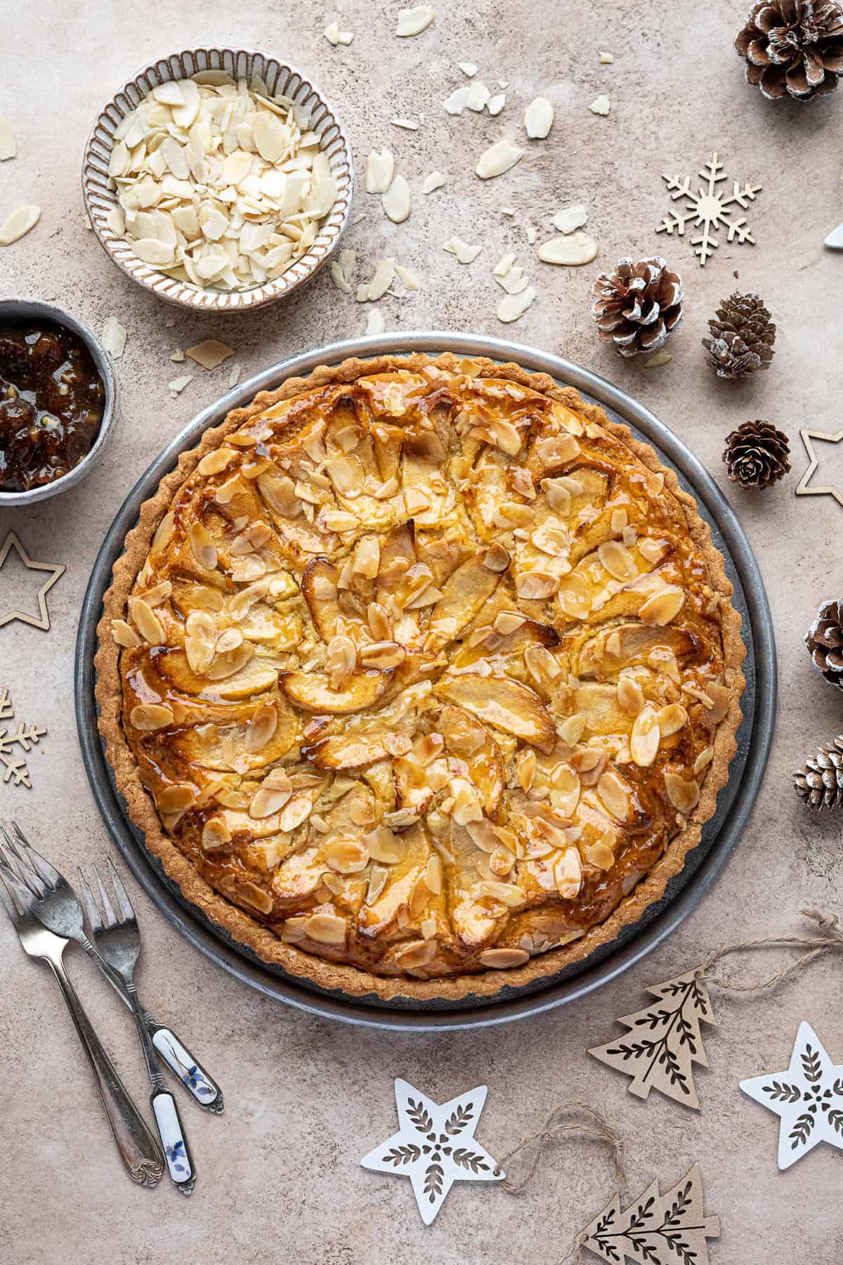 Vegan mincemeat frangipane tart on a metal plate surrounded by pinecones, Christmas decorations and bowls of flaked almonds and mincemeat.