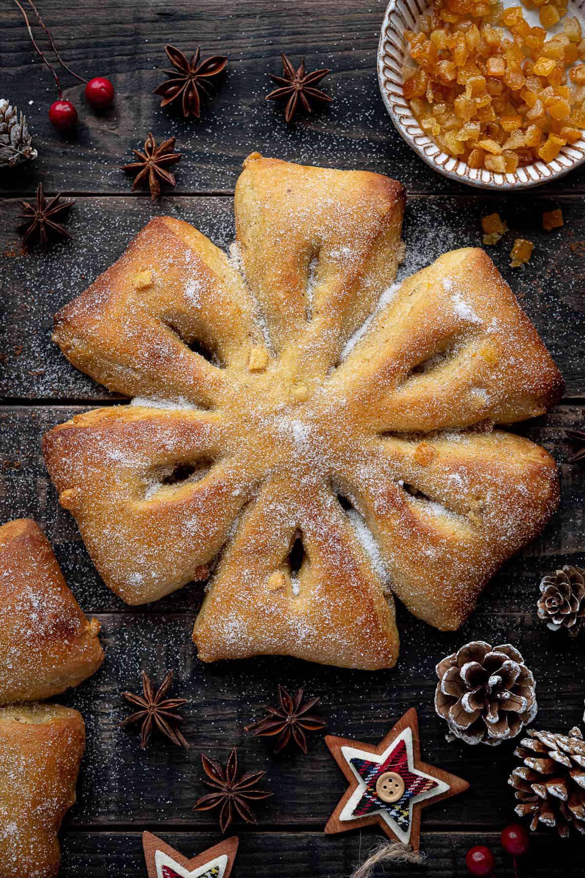 Orange and anise snowflake bread on a wooden background surrounded by Christmas decorations and a bowl of candied peel.