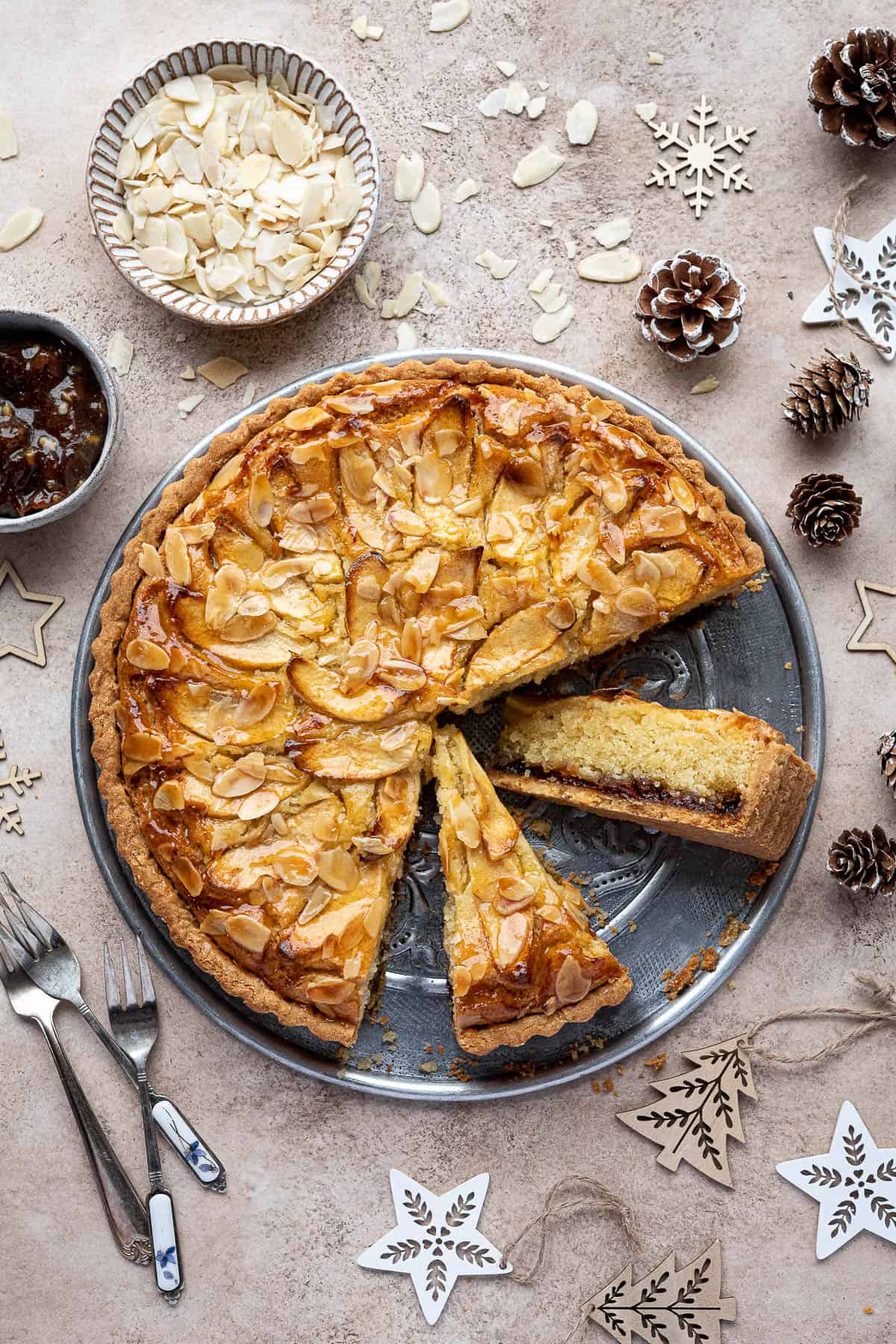 Mincemeat frangipane tart with two slices cut out, surrounded by Christmas decorations an bowls of flaked almonds and mincemeat.