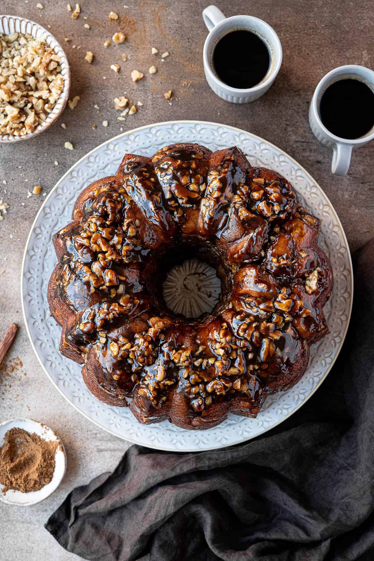 Vegan banana monkey bread  on a white plate with cups of coffee and bowls of walnuts and cinnamon.