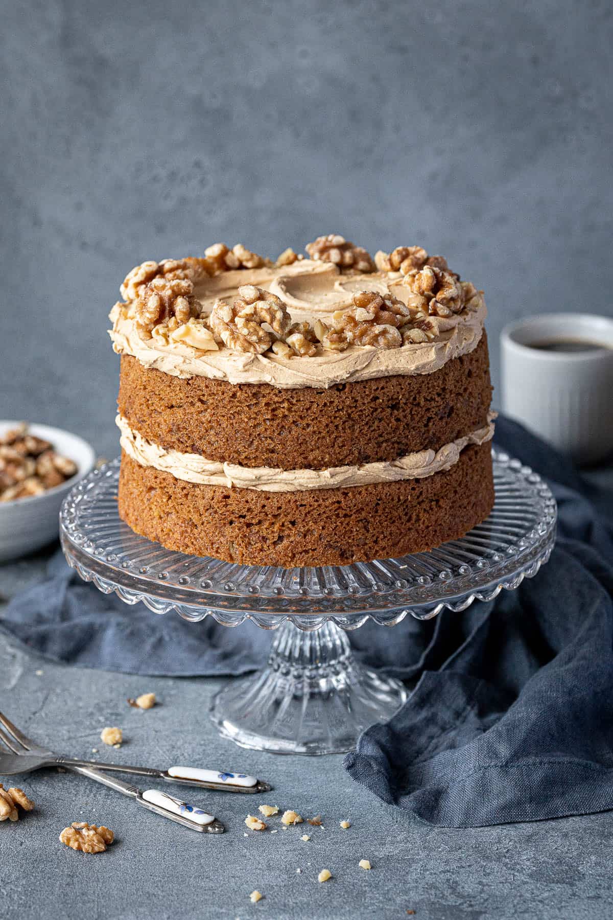 Gluten-free vegan coffee and walnut cake on a glass cake stand with a bowl of walnuts and cups of coffee.