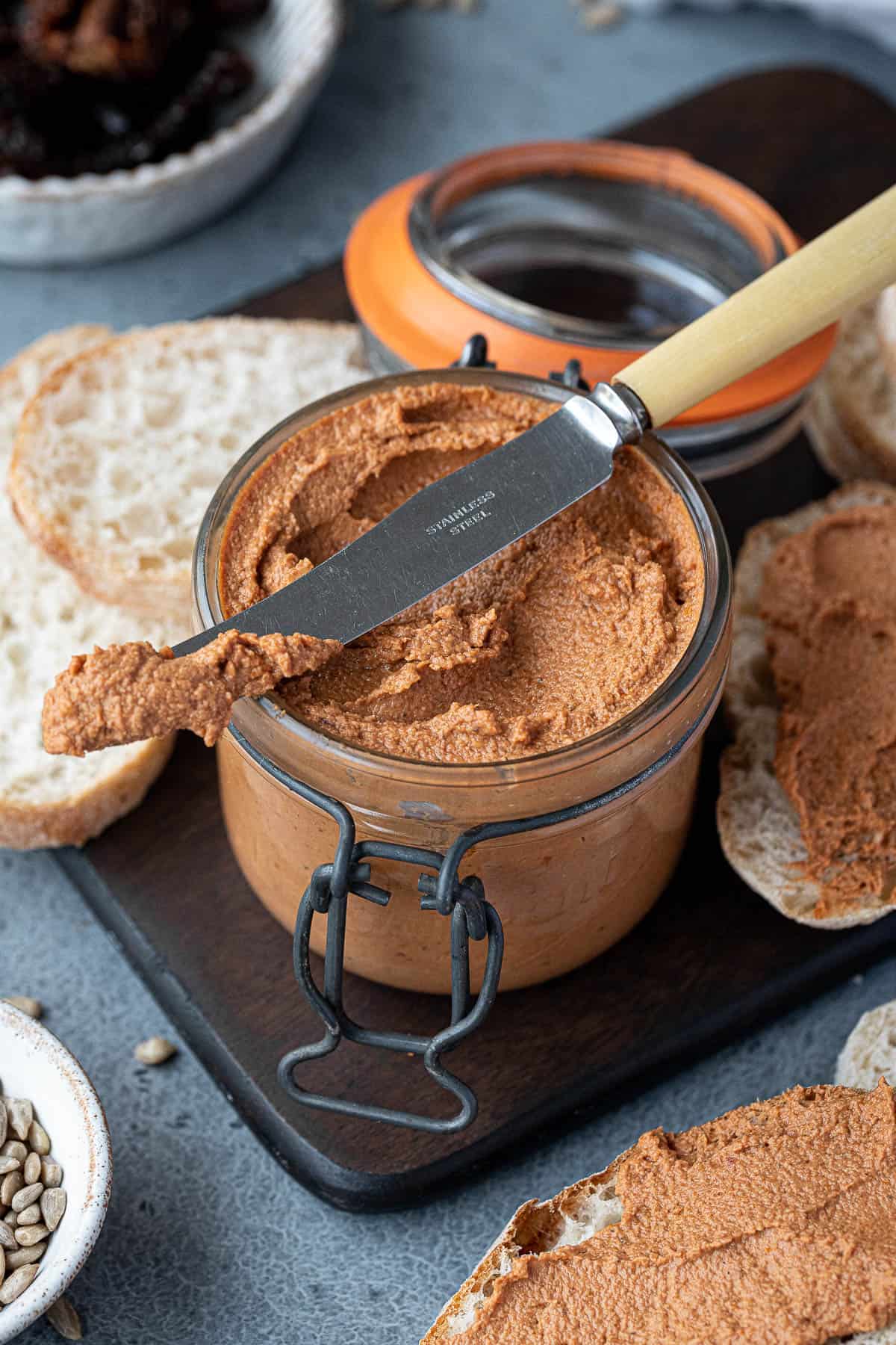 A jar of tomato sunflower seed pate on a  wooden board with a knife and slices of bread.